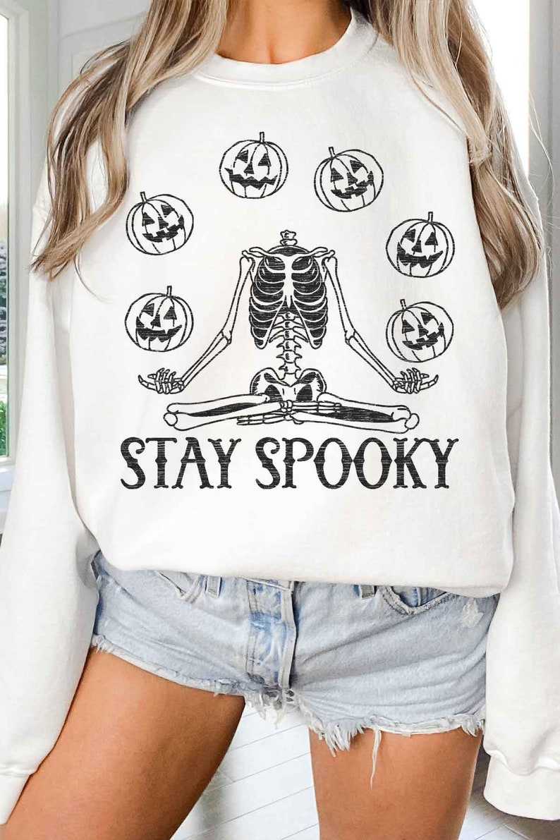 May include: White sweatshirt with a black skeleton meditating with pumpkins around it. The text "STAY SPOOKY" is printed below the skeleton.