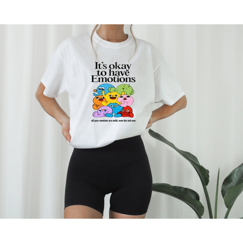 Op de afbeelding: Wit T-shirt met een afbeelding van kleurrijke cartoonfiguren en de tekst "It's okay to have Emotions. All your emotions are valid, even the sad ones."