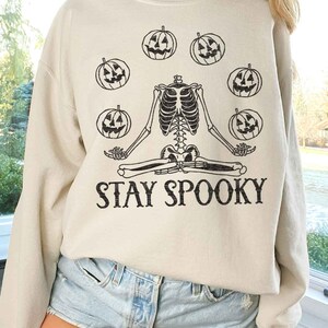 May include: A beige sweatshirt with a skeleton sitting in a yoga pose surrounded by pumpkins. The text "STAY SPOOKY" is printed below the skeleton.