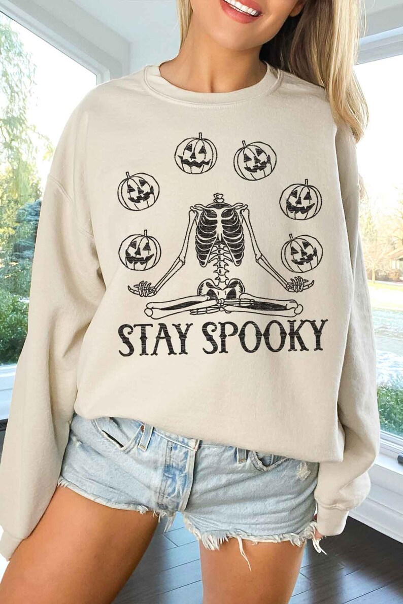 May include: A beige sweatshirt with a skeleton sitting in a yoga pose surrounded by pumpkins. The text "STAY SPOOKY" is printed below the skeleton.