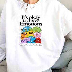 Peut inclure: Sweat-shirt blanc avec un motif de personnages de dessins anim&eacute;s color&eacute;s et le texte "It's okay to have Emotions. All your emotions are valid... even the sad ones."