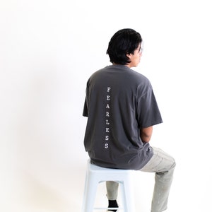 May include: A person wearing a dark gray t-shirt with the word "FEARLESS" printed vertically down the back. The person is sitting on a light blue metal stool.