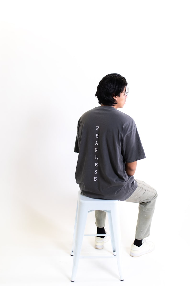May include: A person wearing a dark gray t-shirt with the word "FEARLESS" printed vertically down the back. The person is sitting on a light blue metal stool.
