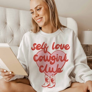 K&ouml;nnte beinhalten: Wei&szlig;er Pullover mit einem rosa Herz-Motiv, auf dem in roten Buchstaben "self love cowgirl club" steht. Zwei rote Cowboystiefel befinden sich am unteren Rand des Herzens.