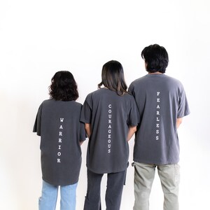May include: Three people wearing gray t-shirts with white text on the back. The text reads "Fearless", "Courageous", and "Warrior".