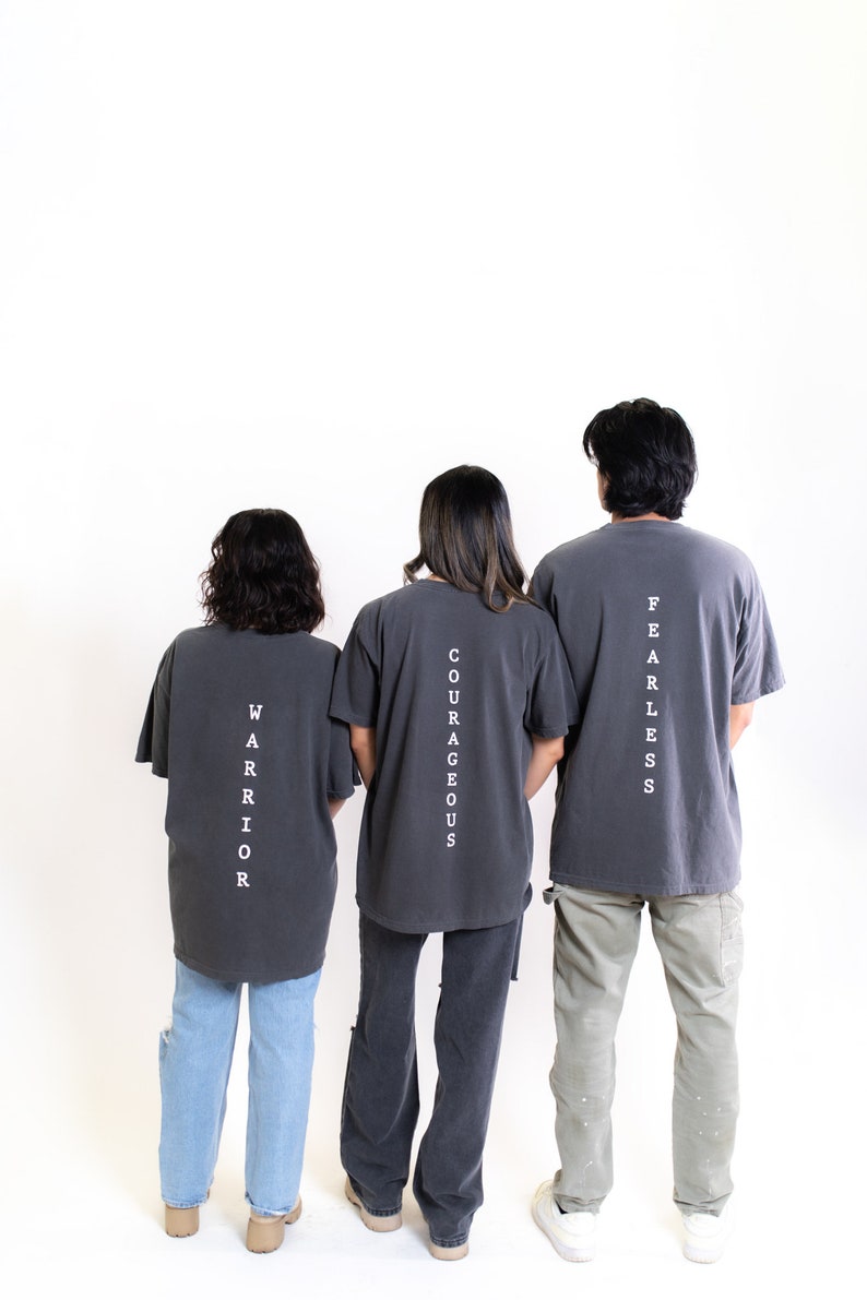 May include: Three people wearing gray t-shirts with white text on the back. The text reads "Fearless", "Courageous", and "Warrior".