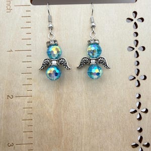 May include: A pair of angel earrings. Each earring features a faceted, iridescent blue bead, silver-colored wings, and a silver-colored hook. The earrings are displayed against a light wood background with a ruler.