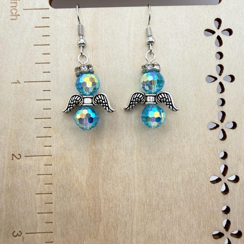 May include: A pair of angel earrings. Each earring features a faceted, iridescent blue bead, silver-colored wings, and a silver-colored hook. The earrings are displayed against a light wood background with a ruler.