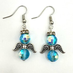 May include: A pair of angel-themed earrings. Each earring features a blue, faceted bead body, silver-colored wings, and a silver-colored hook. The earrings have a small, decorative silver crown above the top bead. The beads have an iridescent finish.