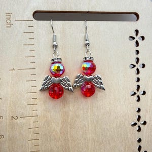 Puede incluir: Un par de pendientes de &aacute;ngel. Cada pendiente presenta una cuenta roja facetada, una cuenta iridiscente m&aacute;s peque&ntilde;a y alas y un coraz&oacute;n de metal plateado. Los pendientes cuelgan de ganchos para la oreja plateados. Sobre un fondo de madera.