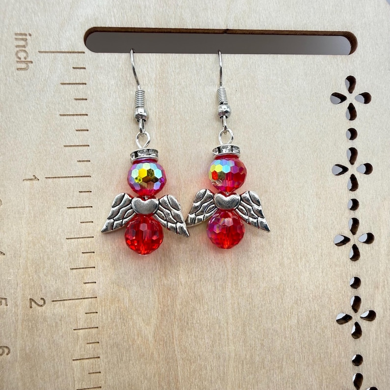 Puede incluir: Un par de pendientes de &aacute;ngel. Cada pendiente presenta una cuenta roja facetada, una cuenta iridiscente m&aacute;s peque&ntilde;a y alas y un coraz&oacute;n de metal plateado. Los pendientes cuelgan de ganchos para la oreja plateados. Sobre un fondo de madera.