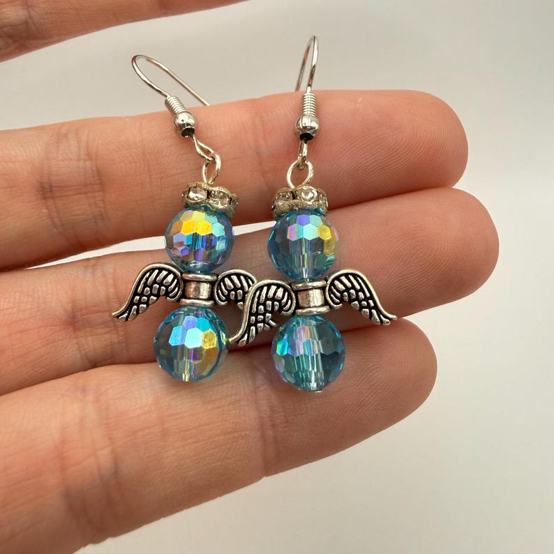 May include: A pair of angel earrings. Each earring features a faceted, iridescent blue bead body, silver-toned wings, and a crown-like top. The earrings are suspended from silver-toned ear wires.