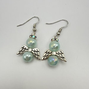 May include: A pair of angel-themed earrings. Each earring features a pearlescent blue bead, silver-colored metal wings, and a decorative top. The earrings have a hook closure. The background is a solid white.