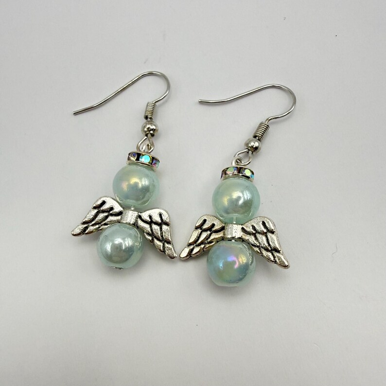 May include: A pair of angel-themed earrings. Each earring features a pearlescent blue bead, silver-colored metal wings, and a decorative top. The earrings have a hook closure. The background is a solid white.