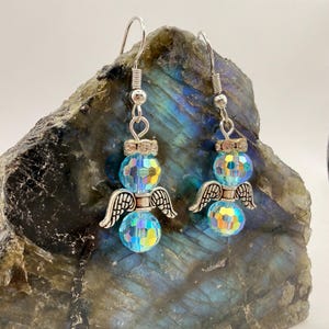 May include: A pair of angel-themed earrings. Each earring features a faceted, iridescent blue crystal bead body, silver-colored wings, and a silver-colored hook. The earrings are designed to dangle and catch the light.