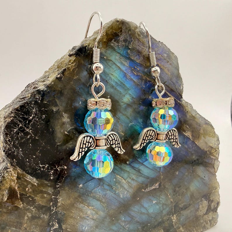 May include: A pair of angel-themed earrings. Each earring features a faceted, iridescent blue crystal bead body, silver-colored wings, and a silver-colored hook. The earrings are designed to dangle and catch the light.