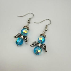 May include: A pair of angel-themed earrings. Each earring features a faceted, iridescent blue bead, a silver-colored crown, and small silver-colored wings. The earrings have silver-colored fish hook closures.