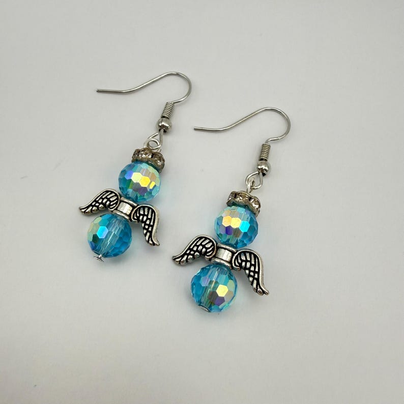 May include: A pair of angel-themed earrings. Each earring features a faceted, iridescent blue bead, a silver-colored crown, and small silver-colored wings. The earrings have silver-colored fish hook closures.