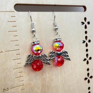 Puede incluir: Un par de pendientes colgantes con dise&ntilde;os de &aacute;ngeles. Cada pendiente tiene una cuenta facetada roja, un dije de ala plateada y una cuenta iridiscente m&aacute;s peque&ntilde;a. Los pendientes cuelgan de ganchos plateados.
