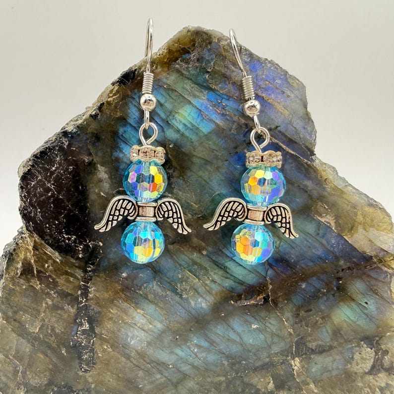 May include: A pair of dangle earrings featuring iridescent blue and yellow faceted beads. Each earring has a silver-toned angel wing charm and a decorative silver-toned top. The earrings are displayed on a textured, multi-colored stone.