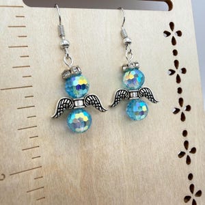 May include: A pair of dangle earrings featuring a silver angel design. Each earring has a faceted, light blue bead, silver wings, and a decorative silver crown. The earrings hang from silver fish hook ear wires.