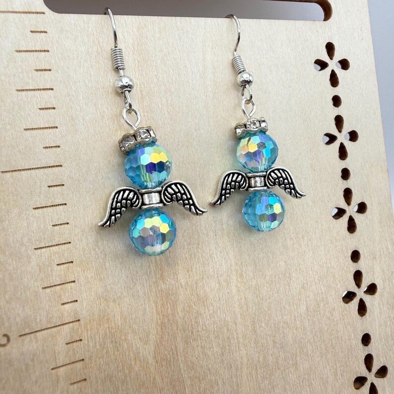 May include: A pair of dangle earrings featuring a silver angel design. Each earring has a faceted, light blue bead, silver wings, and a decorative silver crown. The earrings hang from silver fish hook ear wires.