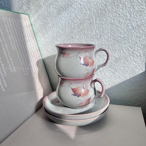May include: Two stacked teacups with floral designs in shades of pink and brown. The cups are on a white saucer with a matching floral design.