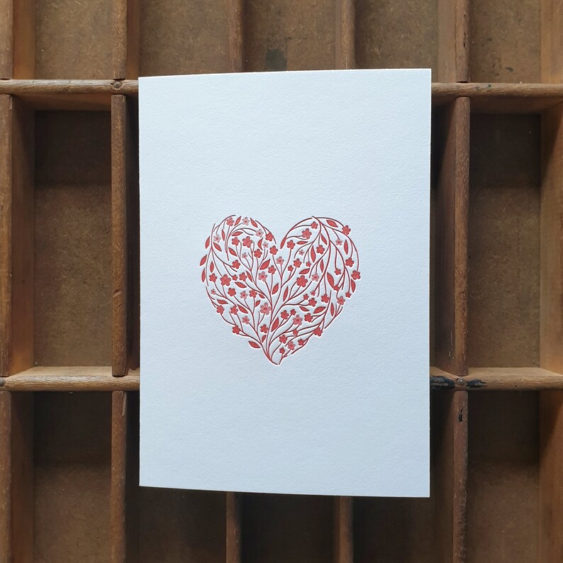 Puede incluir: Una tarjeta blanca con un dise&ntilde;o en forma de coraz&oacute;n rojo hecho de peque&ntilde;as flores y hojas.