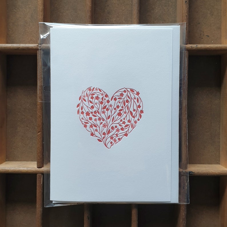 Puede incluir: Una tarjeta blanca con un dise&ntilde;o de coraz&oacute;n floral rojo. El coraz&oacute;n est&aacute; formado por peque&ntilde;as flores rojas y hojas.