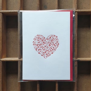 Puede incluir: Una tarjeta blanca con un dise&ntilde;o en forma de coraz&oacute;n rojo hecho de flores y hojas. La tarjeta est&aacute; empaquetada en una funda de pl&aacute;stico transparente con un sobre rojo.