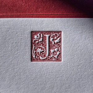 May include: A white letter "J" in a red, ornate, square frame with floral designs.