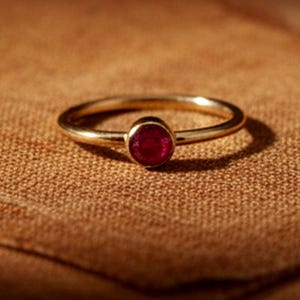 Könnte beinhalten: Ein goldener Ring mit einem kleinen, runden, roten Edelstein. Der Ring liegt auf einem braunen Stoff, wodurch ein Kontrast zwischen dem glänzenden Metall und dem strukturierten Hintergrund entsteht. Der Ring ist schmal und filigran.