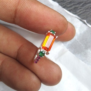 May include: A silver ring featuring an orange rectangular gemstone, surrounded by smaller green and pink gemstones.
