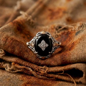 May include: An oval-shaped ring featuring a smooth black stone set in a silver-toned metal band with intricate filigree detailing. A small, four-pointed star-shaped cluster of clear, sparkling stones is centered on the black oval surface.