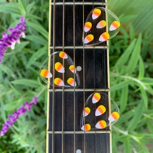 May include: Three guitar picks with a candy corn design. The picks are clear and have yellow, orange, and white candy corn shapes on them. The picks are on a guitar neck.