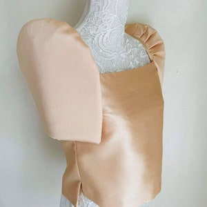May include: A peach-colored satin top with large, puffed sleeves. The square neckline and tailored bodice create a structured silhouette. The top is displayed on a white mannequin with a lace overlay, showcasing the garment's design and texture.
