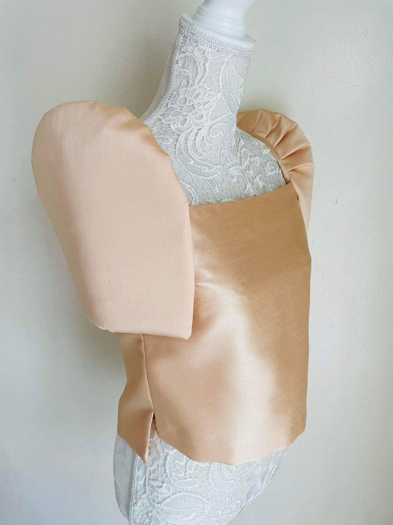 May include: A peach-colored satin top with large, puffed sleeves. The square neckline and tailored bodice create a structured silhouette. The top is displayed on a white mannequin with a lace overlay, showcasing the garment's design and texture.