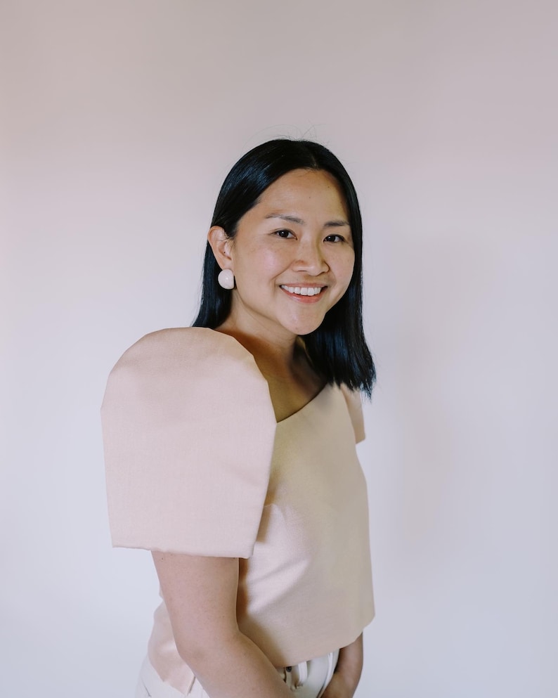 May include: A woman wearing a light peach-colored top with a unique one-shoulder design and a large, puffed sleeve. She is also wearing matching peach-colored earrings. The background is a plain, light peach color.