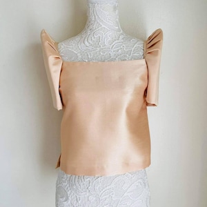 May include: A peach-colored, satin top with square neckline and short, puffed sleeves. The top is displayed on a white mannequin with a lace-covered torso. The fabric has a subtle sheen, suggesting a formal or special occasion garment.