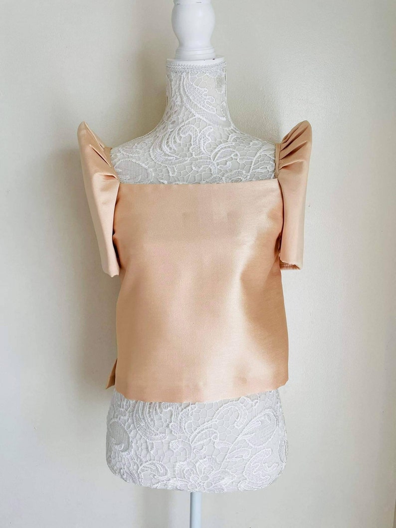 May include: A peach-colored, satin top with square neckline and short, puffed sleeves. The top is displayed on a white mannequin with a lace-covered torso. The fabric has a subtle sheen, suggesting a formal or special occasion garment.