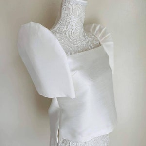 May include: A white, square-neck top with large, puffed sleeves. The top is made of a smooth, satin-like fabric and is displayed on a white mannequin. The mannequin's neck is adorned with delicate lace.