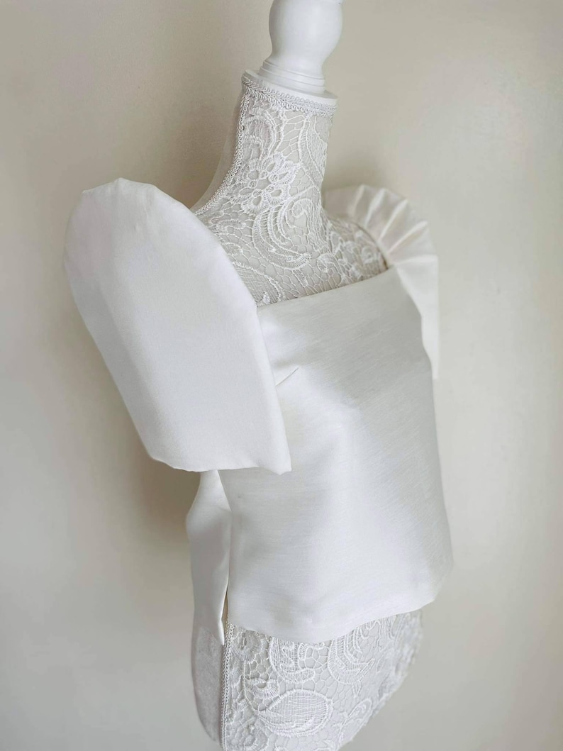 May include: A white, square-neck top with large, puffed sleeves. The top is made of a smooth, satin-like fabric and is displayed on a white mannequin. The mannequin's neck is adorned with delicate lace.