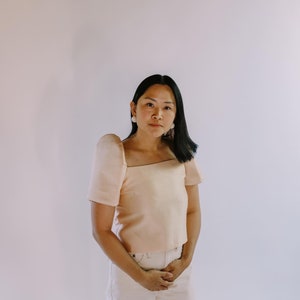 May include: A peach-colored, square-neck top with short sleeves and matching light-colored pants. The top has a structured design, and the outfit is worn by a person with dark hair and white earrings. The background is a plain, light-colored wall.