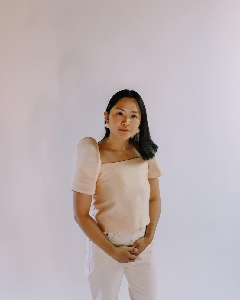 May include: A peach-colored, square-neck top with short sleeves and matching light-colored pants. The top has a structured design, and the outfit is worn by a person with dark hair and white earrings. The background is a plain, light-colored wall.