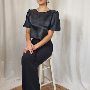 Puede incluir: Una mujer con una blusa de satén negro con mangas abullonadas y pantalones negros de pierna ancha. Está sentada en un taburete blanco frente a un fondo blanco.