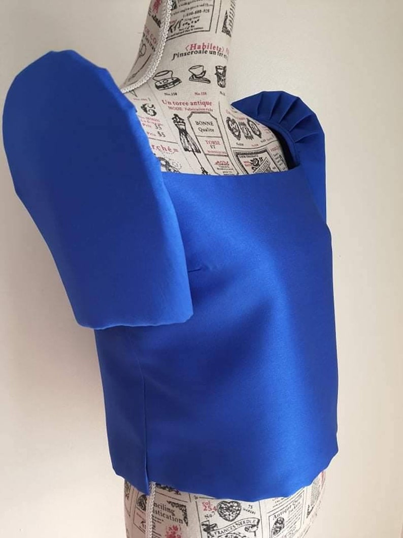 May include: A vibrant blue top with a square neckline and puffed sleeves. The top is made of a smooth, shiny fabric and is displayed on a mannequin. The design features a structured silhouette, showcasing a bold and elegant style.