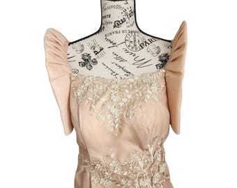 May include: A light brown dress with a fitted bodice and a flowing skirt. The dress features delicate lace detailing and a floral pattern. The dress is on a mannequin with a black stand.