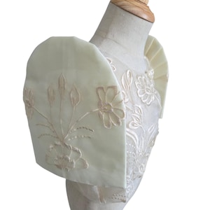 May include: Off-white, embroidered formal top with floral and leaf patterns. The top features a structured shoulder design with a soft, light green hue. The embroidery is in a complementary light beige color, creating a delicate and elegant look.