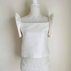 May include: A white satin top with short, puffed sleeves and a square neckline. The top is displayed on a white mannequin with a lace-covered torso. The overall style is elegant and formal, suitable for special occasions.