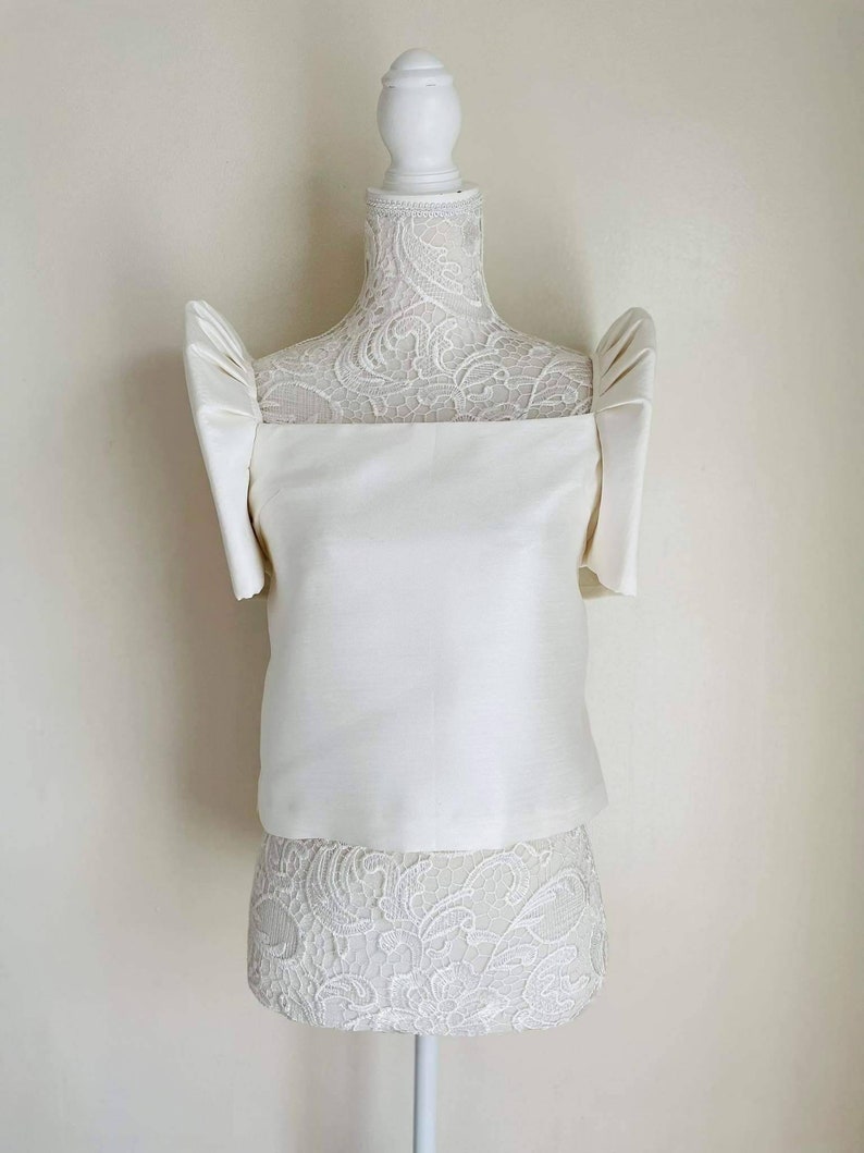 May include: A white satin top with short, puffed sleeves and a square neckline. The top is displayed on a white mannequin with a lace-covered torso. The overall style is elegant and formal, suitable for special occasions.